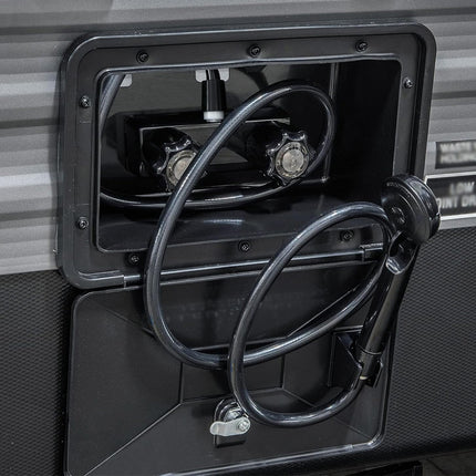 Close-up of a black outdoor shower attachment with a flexible hose and handheld head, mounted inside a recessed compartment on a gray vehicle or equipment surface.