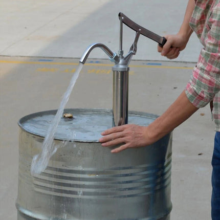 Person using a hand pump to fill a metal barrel with water on a concrete surface.
