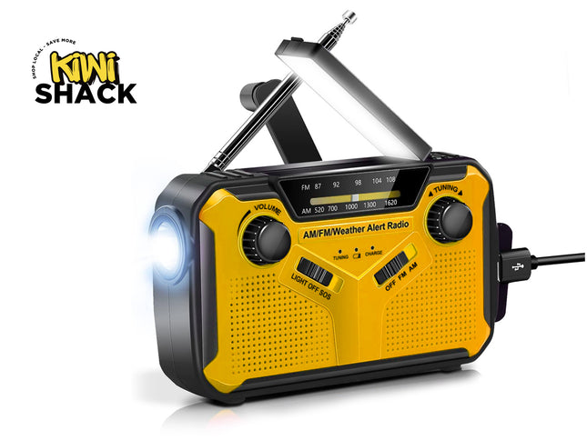 Yellow and black emergency survival solar/hand-crank weather radio featuring a side-mounted bright LED flashlight emitting light beam, an elevated flip-up reading/solar light panel with telescopic antenna, volume and tuning dials, frequency display scale ("AM/FM/Weather Alert Radio"), band/light switches, USB charging cable plugged into the side port, and the Kiwi Shack logo watermark.