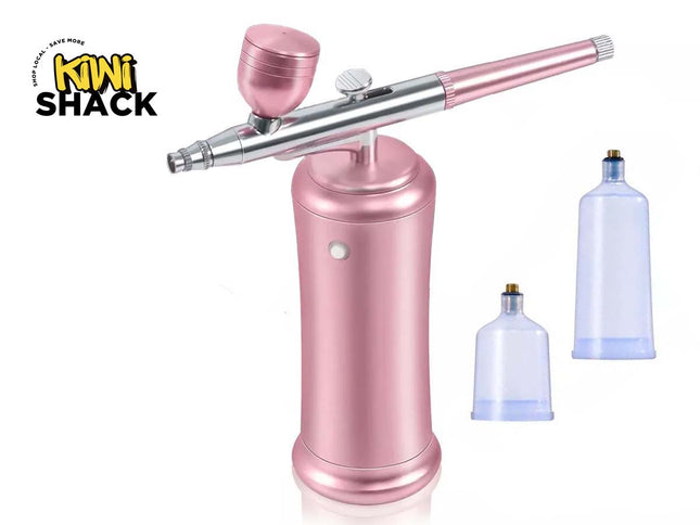 A pink and silver airbrush pen tool with a matching pink cap, shown on a white background with two translucent blue plastic cups. The Kini Shack logo is visible in the top-left corner.