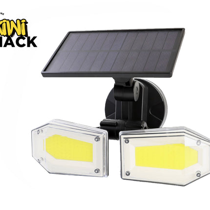 A dual-head solar-powered LED floodlight with bright COB panels enclosed in clear protective covers, mounted on an adjustable black bracket beneath an attached photovoltaic solar panel against a white background, featuring the "Kiwi Shack" logo in the upper left corner.