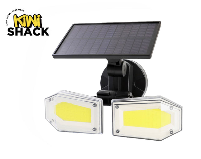 A dual-head solar-powered LED floodlight with bright COB panels enclosed in clear protective covers, mounted on an adjustable black bracket beneath an attached photovoltaic solar panel against a white background, featuring the "Kiwi Shack" logo in the upper left corner.