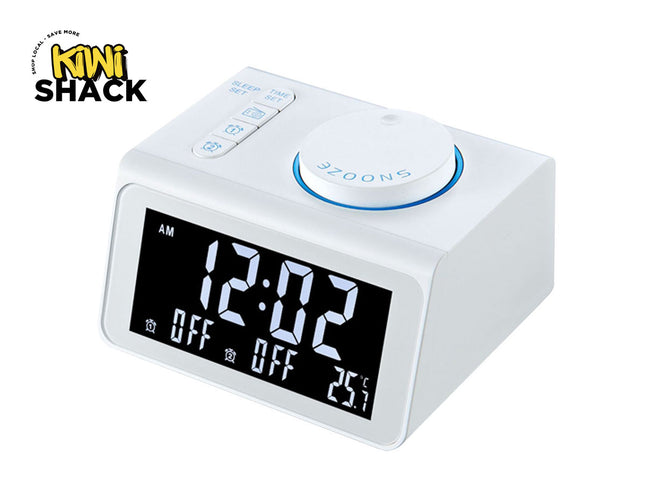 A modern white digital alarm clock with a large LED screen displaying the time, alarm status, and temperature, featuring top function buttons and a large circular snooze dial, with a "Kiwi Shack" logo in the top left corner.