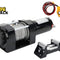 Black electric winch unit with a cylindrical motor housing, steel cable wound on the drum, clutch release knob, tethered cable loop with wired yellow handheld remote control (two push buttons held by a hand), separate black mounting fairlead bracket with four cylindrical steel rollers, and the Kiwi Shack logo watermark.