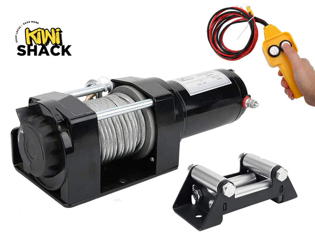 Black electric winch unit with a cylindrical motor housing, steel cable wound on the drum, clutch release knob, tethered cable loop with wired yellow handheld remote control (two push buttons held by a hand), separate black mounting fairlead bracket with four cylindrical steel rollers, and the Kiwi Shack logo watermark.