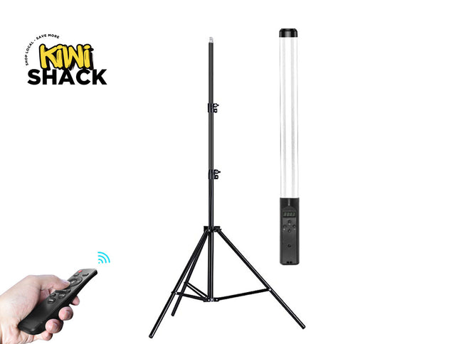 A black tripod light stand with a tall cylindrical lamp attached, shown on a white background. A hand holding a black remote control appears in the lower left, and the Kiwi Shack logo is in the upper left.