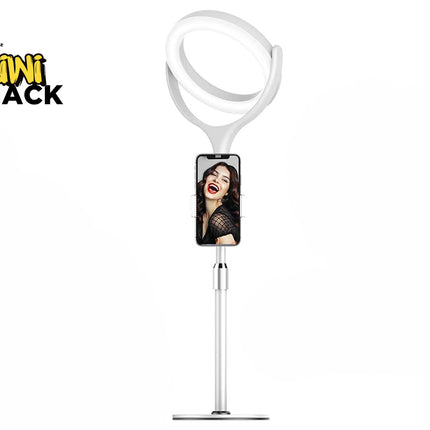 A white desktop ring light stand featuring an adjustable telescopic pole, a phone holder displaying a woman smiling on the screen, and a lit ring light at the top, with a "Kiwi Shack" logo in the top left corner.