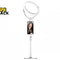 A white desktop ring light stand featuring an adjustable telescopic pole, a phone holder displaying a woman smiling on the screen, and a lit ring light at the top, with a "Kiwi Shack" logo in the top left corner.