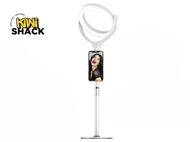 A white desktop ring light stand featuring an adjustable telescopic pole, a phone holder displaying a woman smiling on the screen, and a lit ring light at the top, with a "Kiwi Shack" logo in the top left corner.