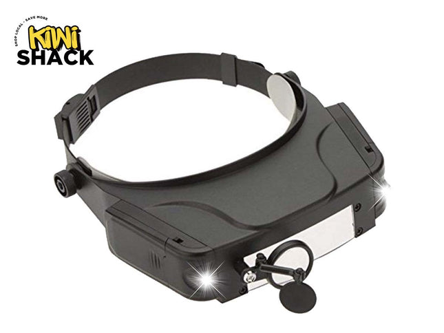 A black magnifying headlamp with an adjustable headband, front light panel, and side adjustment knobs, shown on a white background with a Kiwi Shack logo in the top-left corner.