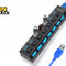 A black multi-port USB hub with individual power switches and blue LED indicators, with two USB flash drives plugged into it and a blue USB cable extending outward, set against a white background with the "Kiwi Shack" logo in the top left corner.