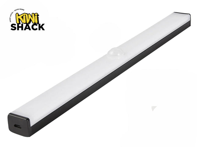 A slim, elongated LED under-cabinet light bar with a black base and a frosted white cover featuring a small motion sensor dome in the center, and a charging port visible on the end cap, with a "Kiwi Shack" logo in the top left corner.