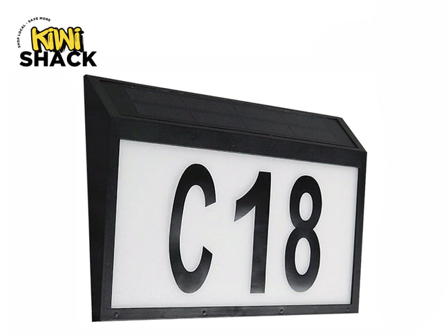A black rectangular solar-powered address sign featuring illuminated white display panels with large bold black characters "C18" and an integrated solar panel on the slanted top surface against a white background, featuring the "Kiwi Shack" logo in the upper left corner.