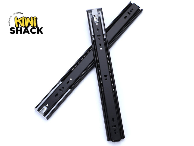 Pair of black heavy-duty full-extension ball-bearing drawer slides crossed over one another, showing mounting hole patterns, metal ball bearings in side raceways, plastic/metal release tabs/stops, and the Kiwi Shack logo watermark