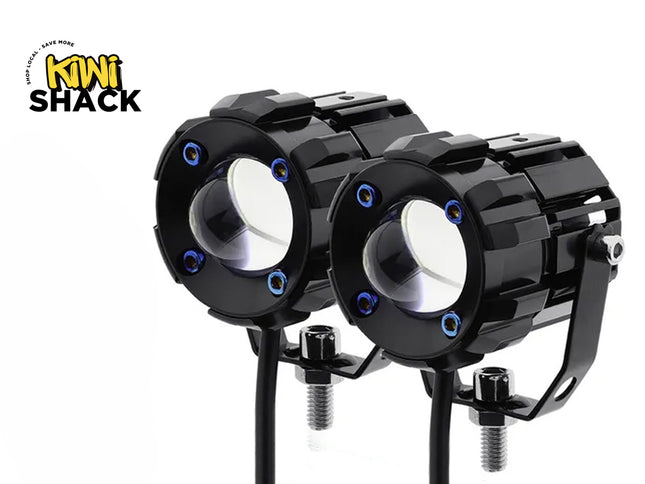 A pair of black round LED projector spotlight pods with U-shaped mounting brackets, threaded bolt bases, and blue accent screws, featuring a "Kiwi Shack" logo in the top left corner.