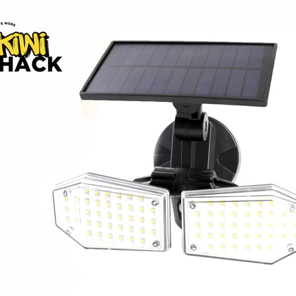 A dual-head solar-powered LED floodlight with individual multi-LED diode panels behind clear protective covers, mounted on an adjustable black bracket beneath an attached photovoltaic solar panel against a white background, featuring the "Kiwi Shack" logo in the upper left corner.