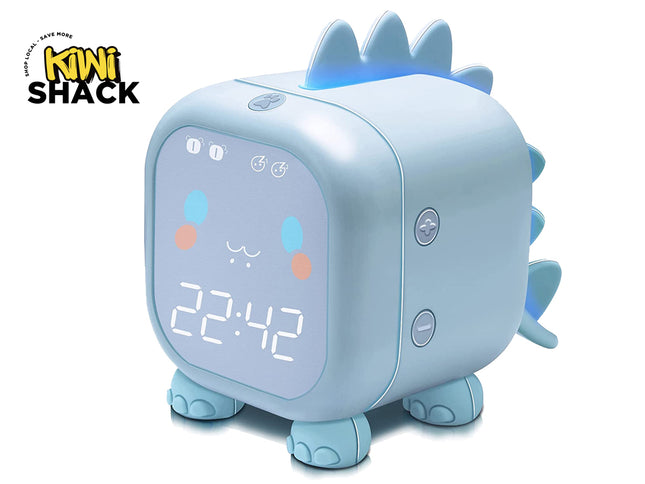 A light blue dinosaur-shaped digital alarm clock featuring a glowing face with a time display, cute facial expressions, soft spikes along the back, and small elephant-like feet against a white background, featuring the "Kiwi Shack" logo in the upper left corner.