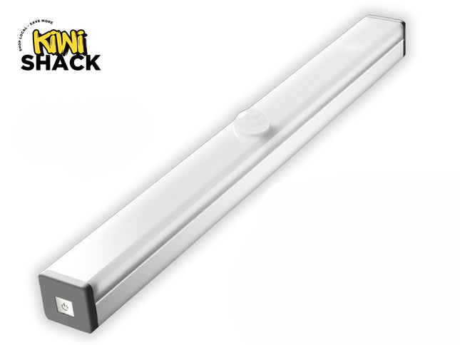 A slim, elongated LED under-cabinet light bar with a silver body and a frosted white cover featuring a circular motion sensor dome on top, along with a power button visible on the gray end cap, featuring a "Kiwi Shack" logo in the top left corner.