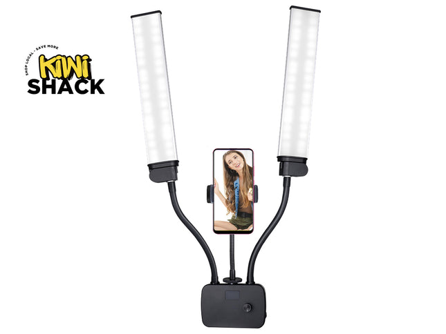 A dual LED light panel selfie stick by Kini Shack, featuring two adjustable flexible arms with bright white light tubes and a central black mounting base. A smartphone is shown in the center holder displaying a selfie video, set against a plain white background with the Kini Shack logo in the top-left corner.