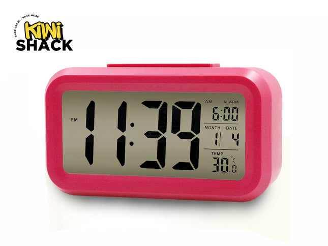 A vibrant pink digital desktop alarm clock featuring a large LCD screen with bold black numbers indicating 11:39 PM, an alarm time set to 6:00, calendar month and date, and a temperature display in Celsius against a white background, featuring the "Kiwi Shack" logo in the upper left corner.