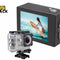 A 4K action camera shown both bare with its textured black housing and back display, and encased in a clear waterproof protective housing mounted on a base, with the screen displaying a surfer on a yellow board against a white background, featuring the "Kiwi Shack" logo in the upper left corner.