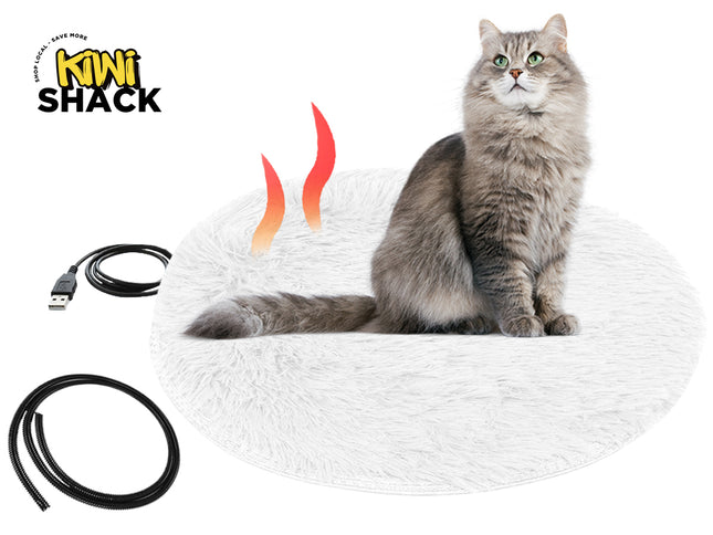 A round white fluffy heated pet bed occupied by a long-haired grey cat, shown alongside a USB power cable and a protective black cord cover against a white background, featuring the "Kiwi Shack" logo in the upper left corner.