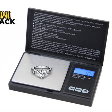 A digital pocket scale weighing a diamond ring on its platform, with operation instructions printed on the inside lid and the "Kiwi Shack" logo in the upper left corner.