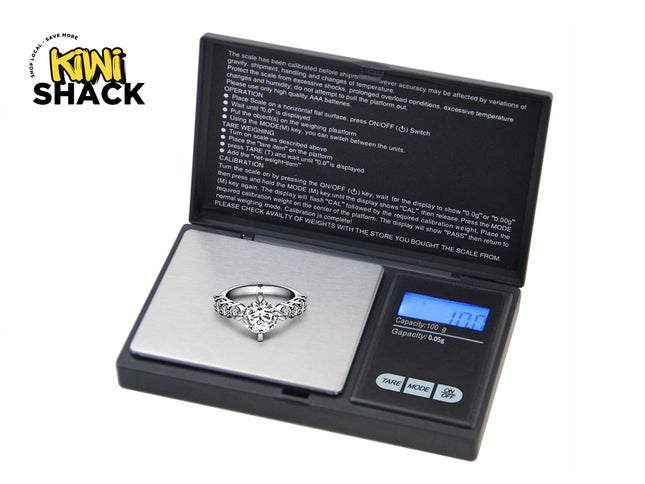 A digital pocket scale weighing a diamond ring on its platform, with operation instructions printed on the inside lid and the "Kiwi Shack" logo in the upper left corner.