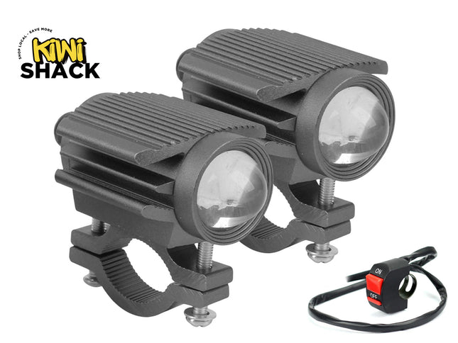 A pair of black auxiliary LED motorcycle spotlight pods with ribbed aluminum heat sinks, projector lenses, handlebar mounting clamps, and an on-off toggle switch, featuring a "Kiwi Shack" logo in the top left corner.