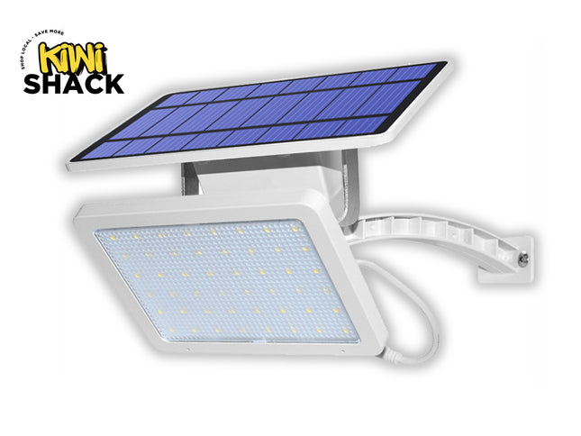 A white solar-powered LED floodlight attached to a curved mounting arm, featuring a rectangular light panel and a separate solar panel mounted on top against a white background, featuring the "Kiwi Shack" logo in the upper left corner.