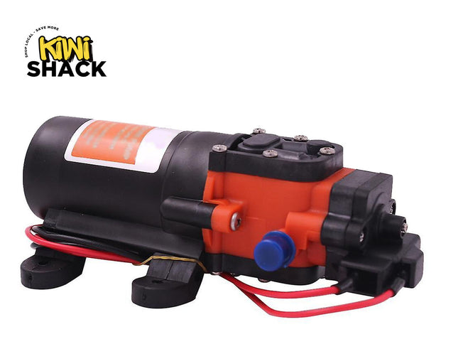 A small electric pump motor with a black cylindrical body, orange casing, black mounting feet, a blue connector cap, and red wires, shown on a white background with the Kiwi Shack logo in the top-left corner.