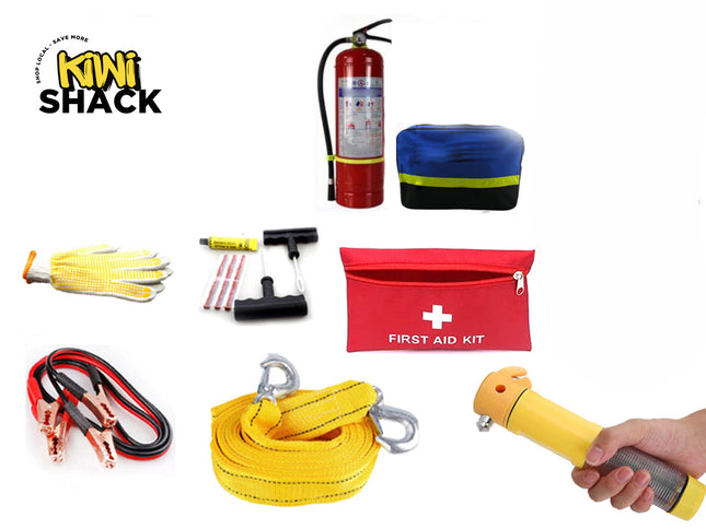 A comprehensive roadside emergency and safety kit laid out against a white background, including a red fire extinguisher, a blue reflective warning triangle case, a red "FIRST AID KIT" bag, heavy-duty jumper cables, a yellow tow strap with hooks, a tire repair set, a pair of work gloves, and a hand holding a yellow emergency escape hammer, with the "Kiwi Shack" logo in the upper left corner.