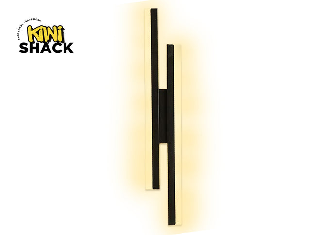 A modern wall-mounted light fixture with two vertical black bars and a central black connector, set against a warm yellow glow. The brand logo 'Kini Shack' is visible in the top-left corner.