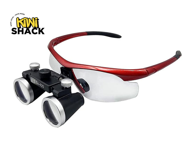 A pair of surgical magnifying glasses with a red frame, clear lenses, and a black double-ocular microscope attachment. The Kiwi Shack logo is visible in the top-left corner.