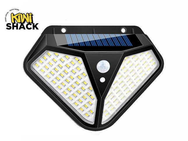 A trapezoidal solar-powered wall light featuring two angled multi-LED panels, a built-in motion sensor on the front center, and a small solar panel integrated into the top section against a white background, featuring the "Kiwi Shack" logo in the upper left corner.