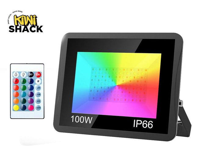 A 100W IP66 waterproof RGB LED floodlight displaying vibrant multi-color illumination on its panel, accompanied by a multi-button remote control, set against a white background. A "Kiwi Shack" logo appears in the top-left corner.