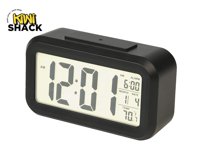 A modern black digital alarm clock with a backlit LCD screen showing time, alarm setting, calendar date, and temperature, featuring the "Kiwi Shack" logo in the top-left corner.