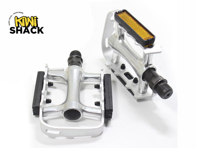 A pair of silver bicycle pedals featuring threaded steel spindles, black rubber platform grips, and integrated orange reflectors against a white background, featuring the "Kiwi Shack" logo in the upper left corner.