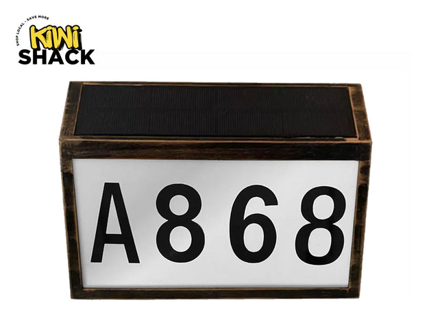 Dark-rimmed rectangular solar-powered LED house number / address plate featuring a dark bronze-finish or wood-grain frame, a black photovoltaic solar panel on top, a white illuminated front panel displaying large black characters "A868", and the Kiwi Shack logo watermark.