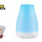 A blue-lit diffuser with a white base and control buttons labeled "LIGHT" and "MIST", next to a hand holding a small remote control, with the "Kiwi Shack" logo in the top left corner.