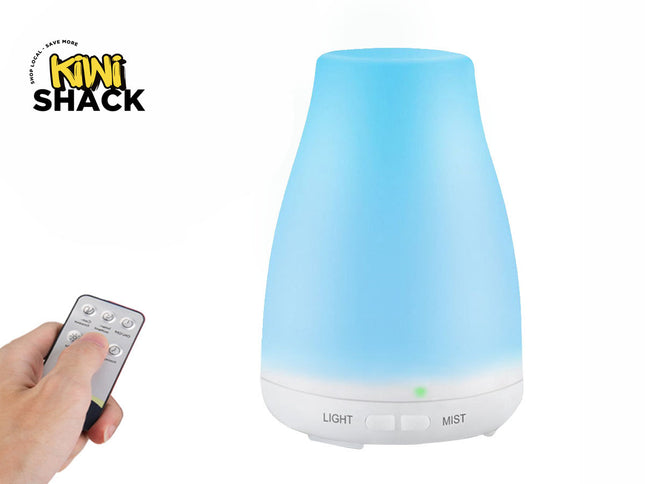 A blue-lit diffuser with a white base and control buttons labeled "LIGHT" and "MIST", next to a hand holding a small remote control, with the "Kiwi Shack" logo in the top left corner.