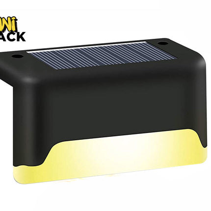 A black solar-powered step light featuring an integrated top solar panel, mounting screw holes, and a glowing warm-toned LED light emission along the bottom edge against a white background, featuring the "Kiwi Shack" logo in the upper left corner.