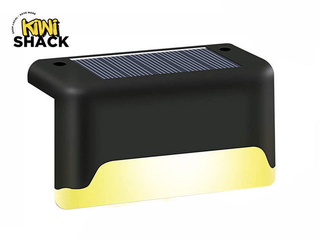A black solar-powered step light featuring an integrated top solar panel, mounting screw holes, and a glowing warm-toned LED light emission along the bottom edge against a white background, featuring the "Kiwi Shack" logo in the upper left corner.