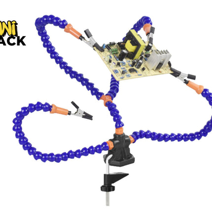 A helping hands soldering tool with flexible blue and orange gooseneck arms featuring alligator clips, secured to a workbench via a black mounting clamp, holding a circuit board in place, with a "Kiwi Shack" logo in the top left corner.