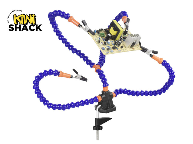 A helping hands soldering tool with flexible blue and orange gooseneck arms featuring alligator clips, secured to a workbench via a black mounting clamp, holding a circuit board in place, with a "Kiwi Shack" logo in the top left corner.