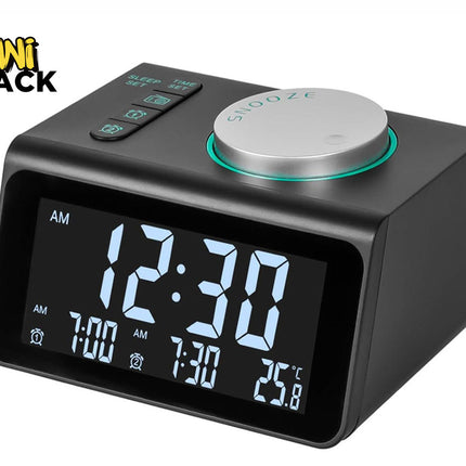 A modern dark grey digital alarm clock with a large LED screen showing the time, dual alarms, and temperature, featuring top function buttons and a large circular silver snooze dial with a turquoise ring, with a "Kiwi Shack" logo in the top left corner.