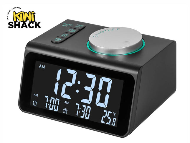 A modern dark grey digital alarm clock with a large LED screen showing the time, dual alarms, and temperature, featuring top function buttons and a large circular silver snooze dial with a turquoise ring, with a "Kiwi Shack" logo in the top left corner.