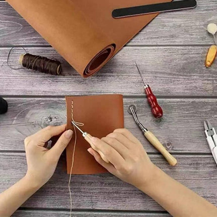 A top-down view of a person's hands stitching a brown leather journal on a gray wooden table, surrounded by leatherworking tools including a spool of thread, a roll of leather, and stitching needles.