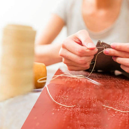 A close-up lifestyle shot of a person's hands sewing a dark brown leather patch with white thread over a red work surface.