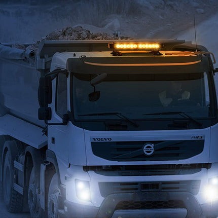 A white Volvo dump truck driving through snow at night, with its headlights on and amber roof-mounted lights illuminated.
